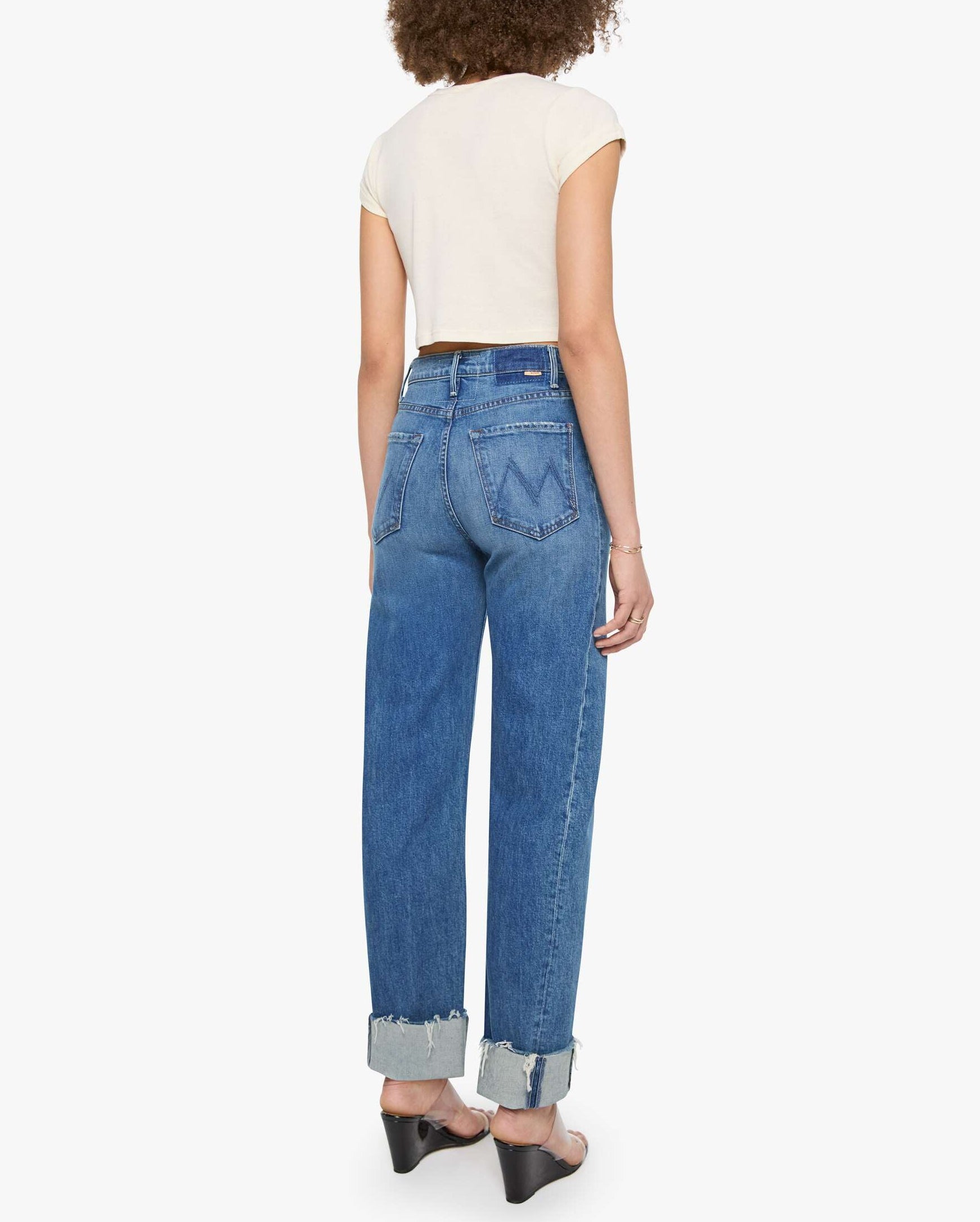 A person stands with their back to the camera, wearing a short-sleeve white top and the Half Pipe Flood Cuff Fray high-waisted, wide-leg jeans by Mother, crafted from SUPERIOR denim with cuffed hems, paired with black heeled sandals.