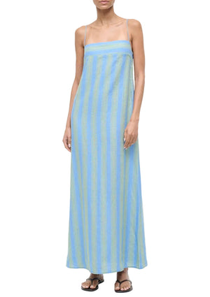 A person wearing the Staud Laura Linen Dress, featuring blue and green stripes and adjustable spaghetti straps, paired with flat sandals, stands against a white background.