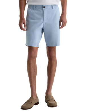 A person wears AG Jeans' Wanderer Shorts in light blue with a tucked-in white shirt and tan loafers, highlighting a tailored style, shown from shoulders to feet.