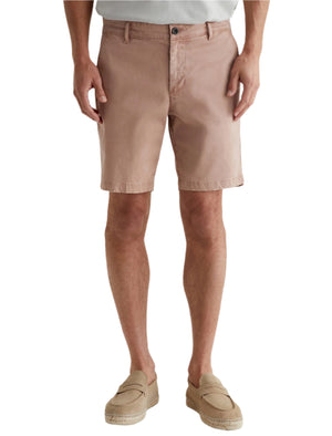 A person in light pink AG Jeans Wanderer Shorts with a tailored look, paired with a white shirt and beige loafers, stands against a plain background.