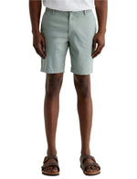 A person wears light green AG Jeans Wanderer Shorts, a white shirt, and brown sandals, standing against a plain background for a tailored look.