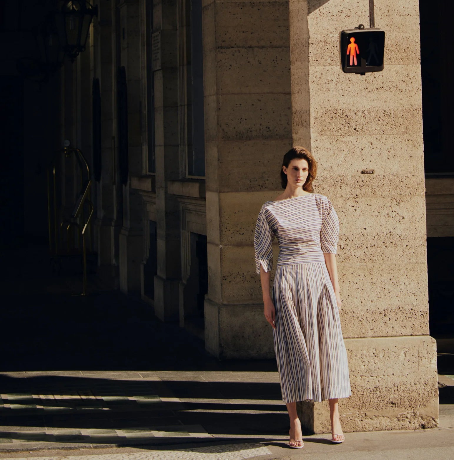 A woman in a Lela Rose Striped Cotton Full Skirt stands on a sidewalk near a stone building under a red pedestrian signal.
