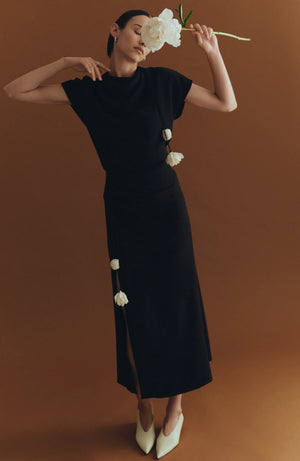 A model poses against a brown background wearing the Lela Rose Drop Flower Ruched Skirt, holding a white flower near their face and pairing the look with white pointed shoes.