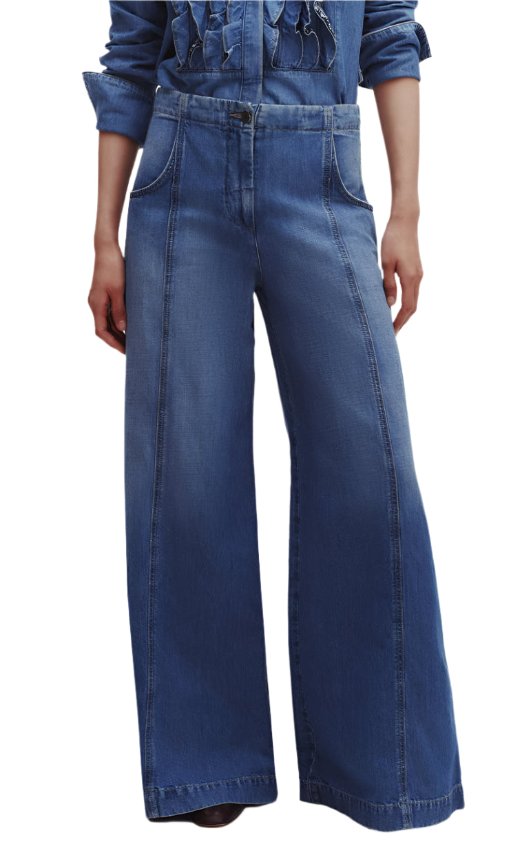 A person wearing TWP's Dancing In The Dark Jeans, featuring blue ultra wide-leg design with front seam details and pockets, paired stylishly with a denim shirt. The relaxed fit crafted from lightweight denim ensures both comfort and chic elegance.