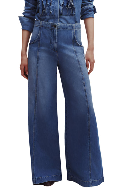 A person wearing TWP's Dancing In The Dark Jeans, featuring blue ultra wide-leg design with front seam details and pockets, paired stylishly with a denim shirt. The relaxed fit crafted from lightweight denim ensures both comfort and chic elegance.