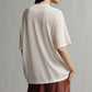 A person with long dark hair is shown from the back, wearing a loose-fitting, short-sleeve white TWP Big Boy Skye Cashmere Tee and brown pants.