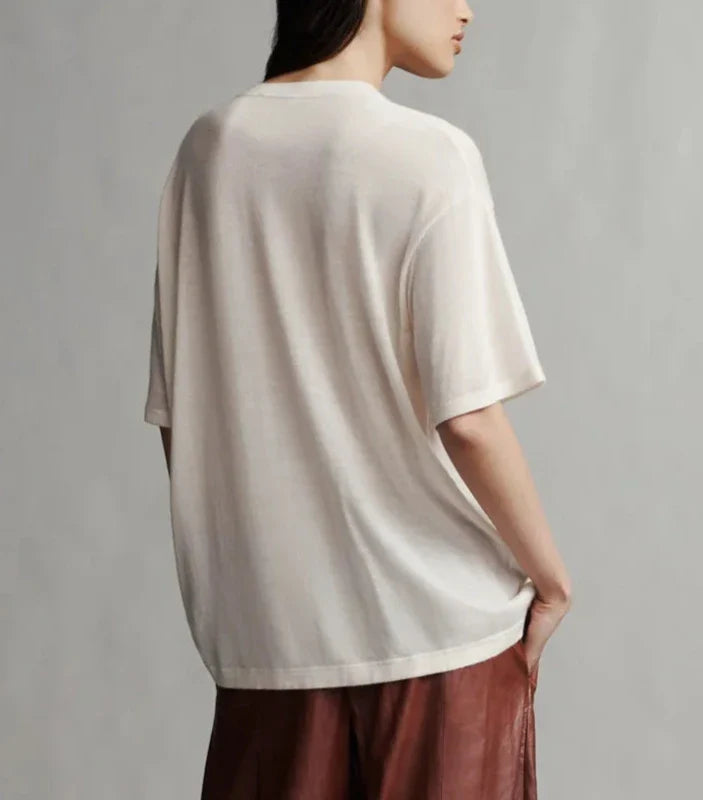 A person with long dark hair is shown from the back, wearing a loose-fitting, short-sleeve white TWP Big Boy Skye Cashmere Tee and brown pants.