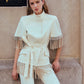 A woman wearing the Lela Rose Stretch Cotton Fringed Cape, styled with an off-white pantsuit featuring fringe details, stands against a stone wall with one hand resting on her thigh as she looks at the camera.