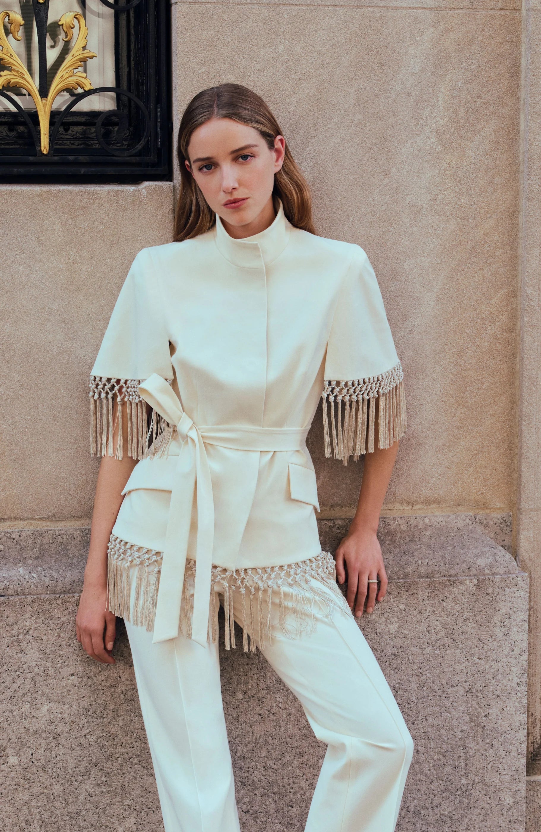 A woman wearing the Lela Rose Stretch Cotton Fringed Cape, styled with an off-white pantsuit featuring fringe details, stands against a stone wall with one hand resting on her thigh as she looks at the camera.
