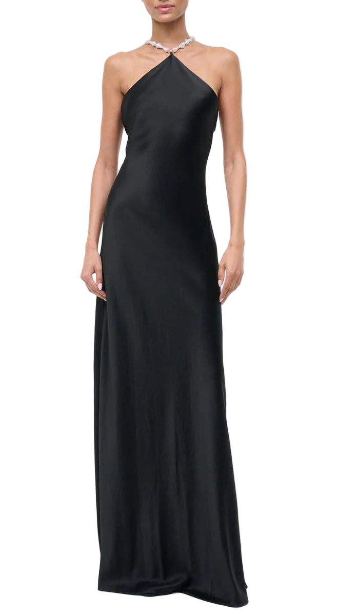 A person wearing the Staud Cadence Dress—a floor-length, sleeveless black satin halter-neck gown with a fitted silhouette—stands against a white background.