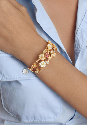 A person in a light blue shirt wears the Anabel Aram Wildflowers Bangle, a hand-painted enamel bracelet featuring gold floral details with white and red accents on their wrist.
