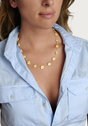 A woman wears the Anabel Aram Wildflowers Station Necklace, a nature-inspired gold piece, over a light blue shirt against a plain background.