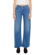 A person wearing the Mother Mid Rise Lasso Sneak—high-waisted, wide-leg blue jeans crafted from SUPERIOR denim—paired with a white crop top and black shoes, standing against a white background.