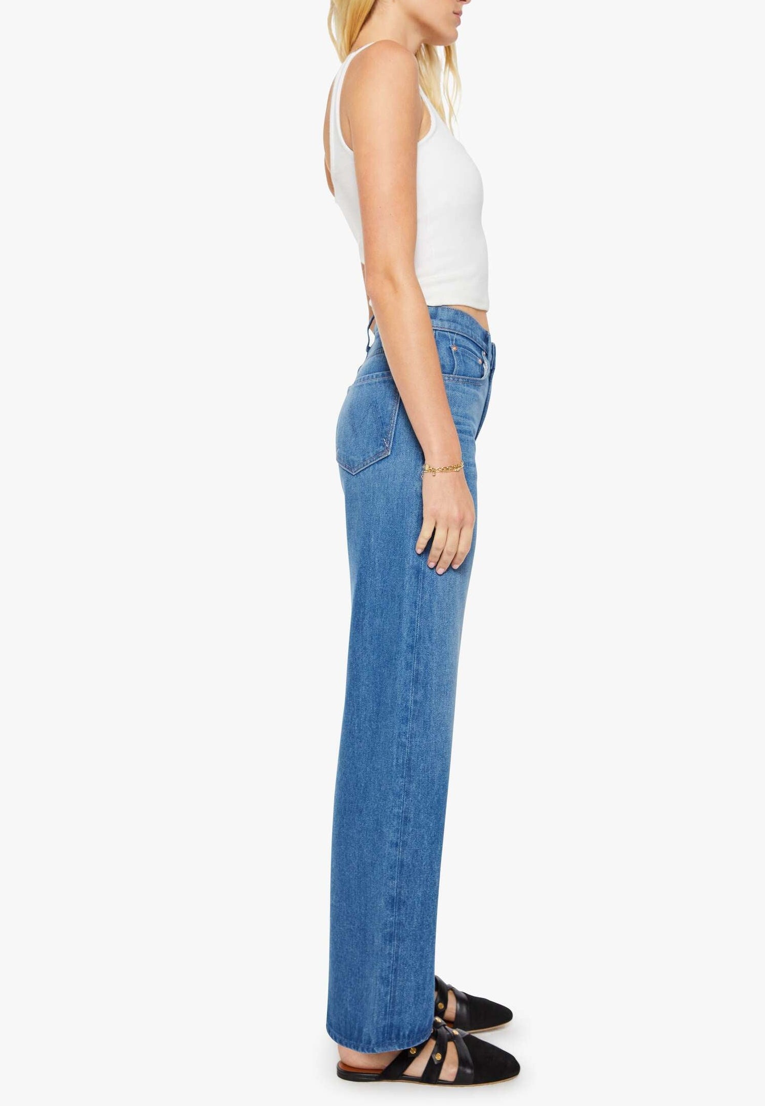 A woman stands in profile against a plain white background, wearing a white sleeveless top, black shoes, and blue Mother Mid Rise Lasso Sneak denim wide-leg pants.