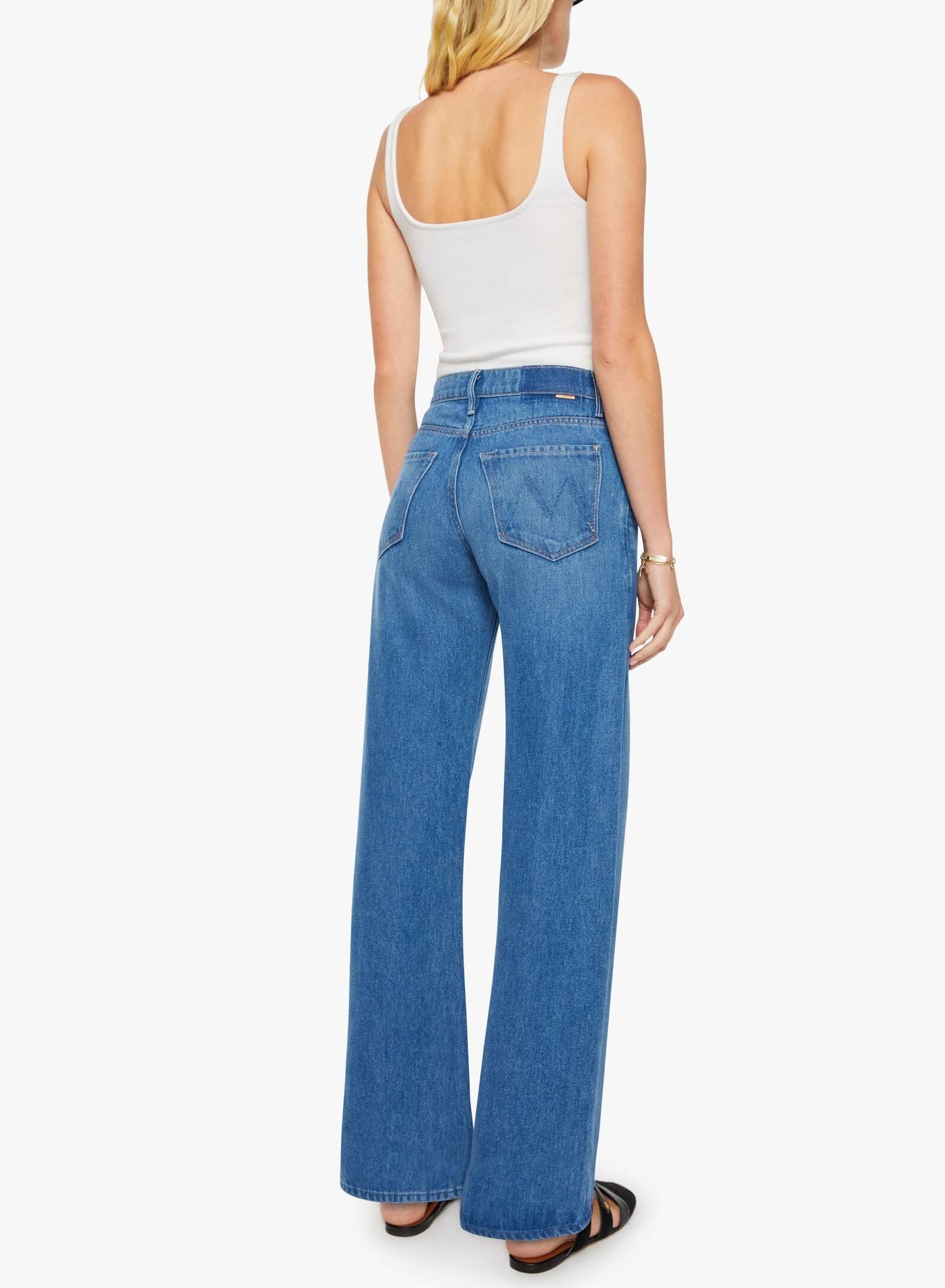 A person stands facing away, wearing a white tank top, the Mid Rise Lasso Sneak high-waisted wide-leg jeans by Mother crafted from SUPERIOR denim, black sandals, and a bracelet, against a plain white background.