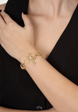 A person in a sleeveless black outfit showcases the Anabel Aram Cheval Chain Bracelet, featuring gold plating and a bar and ring toggle clasp, on their wrist.