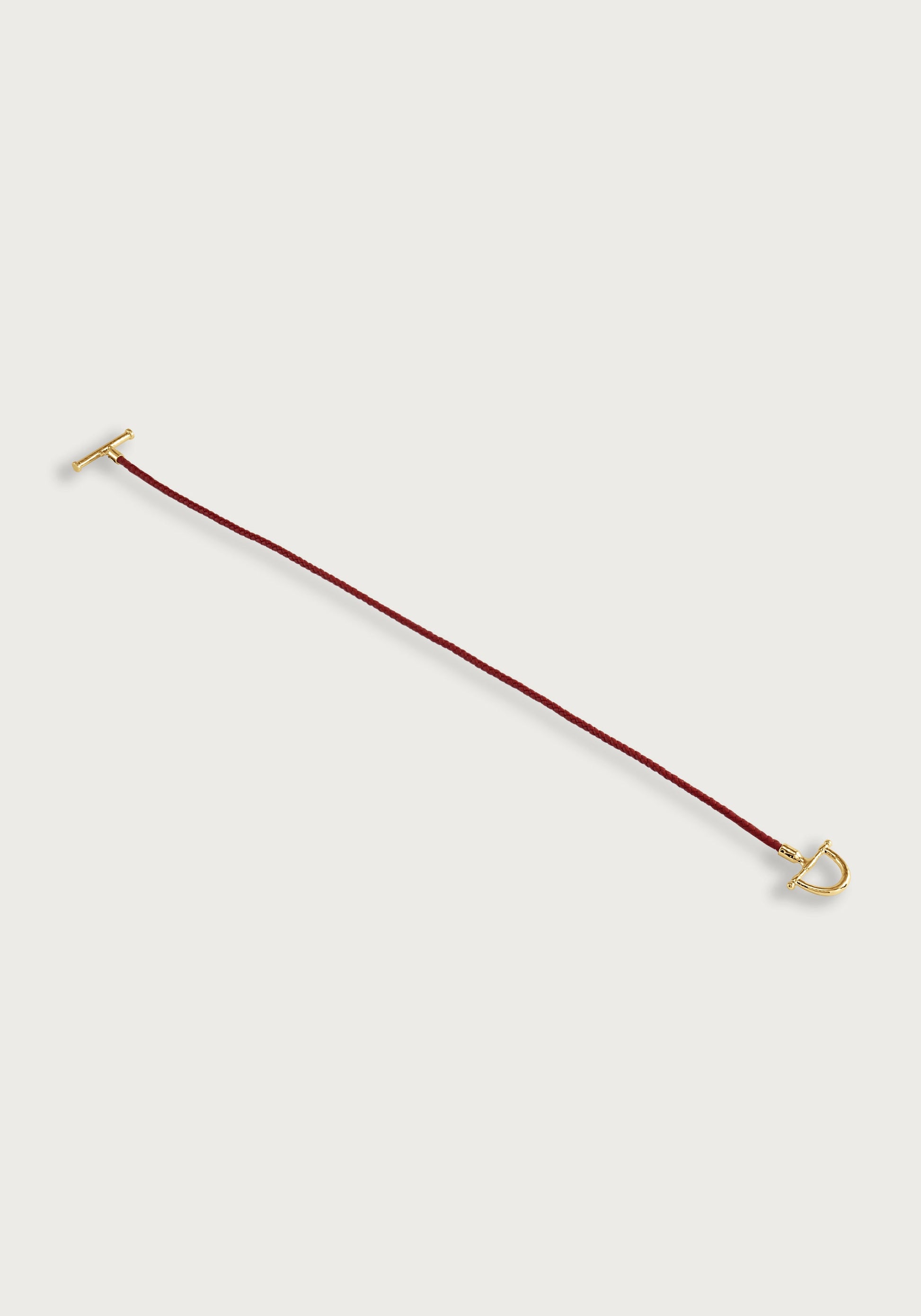 The Cheval Leather Wrap Bracelet by Anabel Aram features a slim red leather cord with gold-tone T-bar and D-shaped clasp, inspired by equestrian style—ideal for daily wear. Shown against a white background.