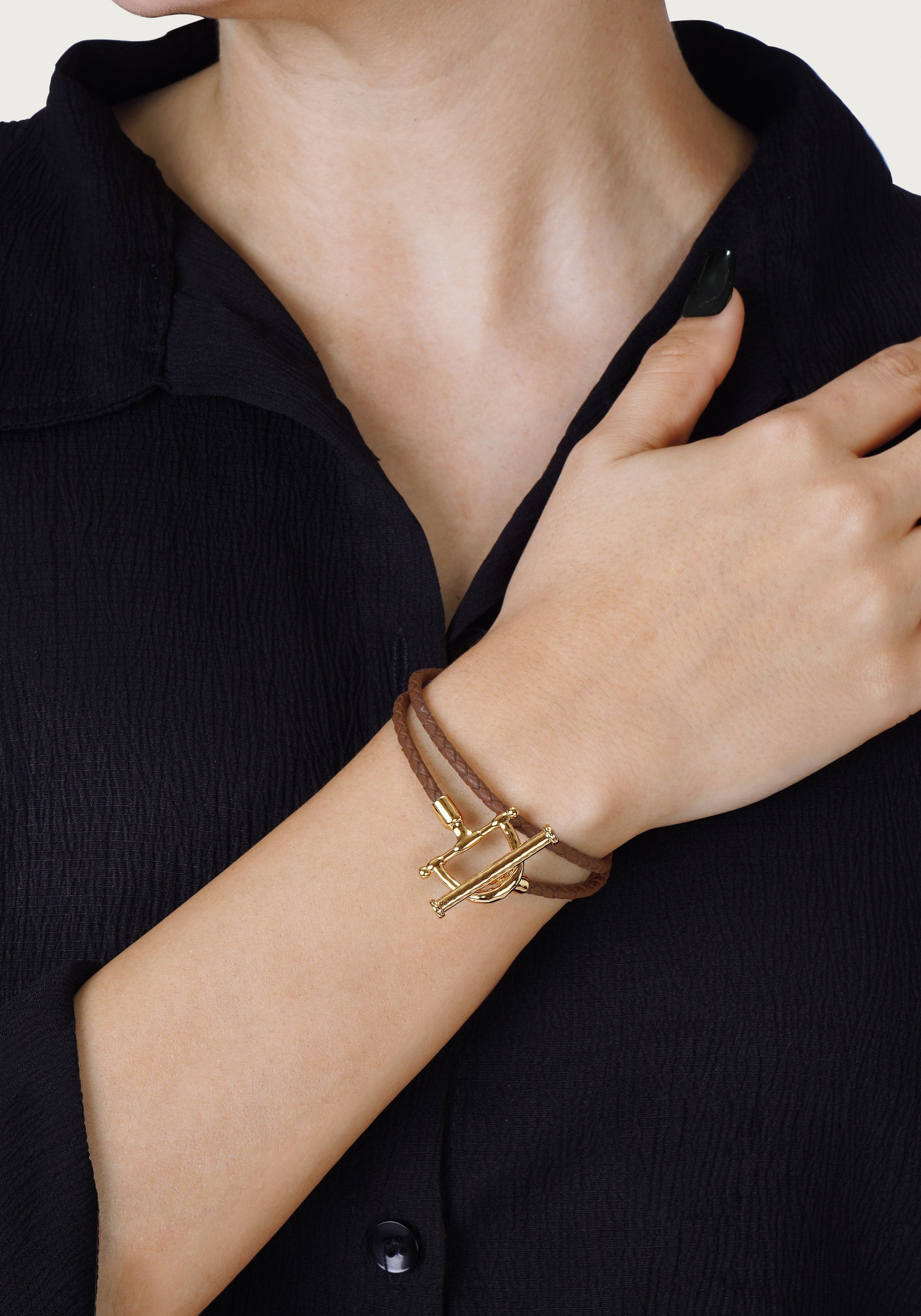 A person wearing a black textured shirt and the Anabel Aram Cheval Leather Wrap Bracelet with gold accents on their wrist, one hand resting on their collarbone.
