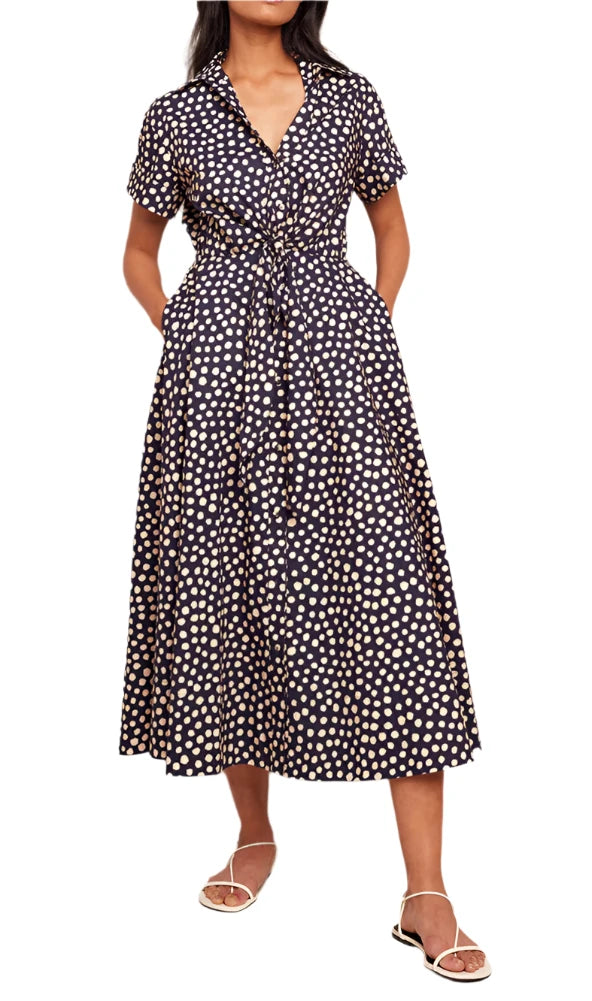 A woman is wearing the Cara Cara Asbury Dress, a navy blue polka dot shirt dress with short sleeves, a self-tie waist, and hands in pockets, paired with white sandals.