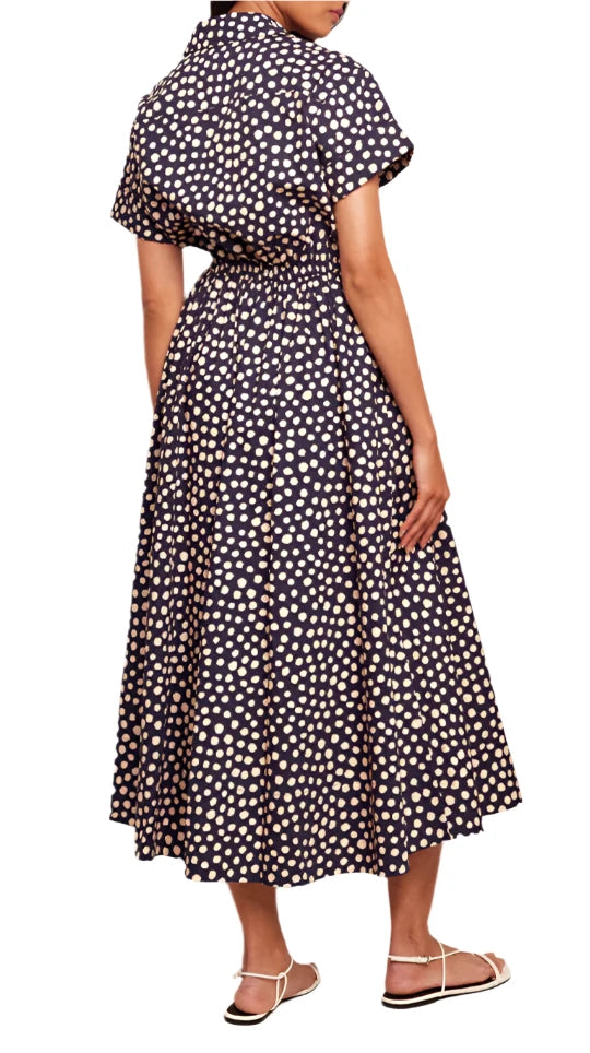 The individual is dressed in the Cara Cara Asbury Dress, featuring a navy blue and white polka dot design with a self-tie waist. Their back is partially turned to highlight the dress's length and pattern while they wear white sandals, exuding a chic Cara Cara look.