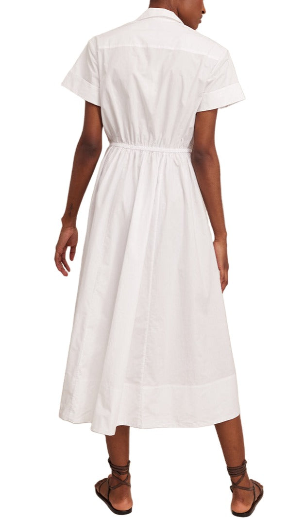 A person wearing the Cara Cara Asbury Dress, a white, short-sleeved cotton poplin shirt dress with a self-tie waist, paired with strappy sandals is standing with their back to the camera.