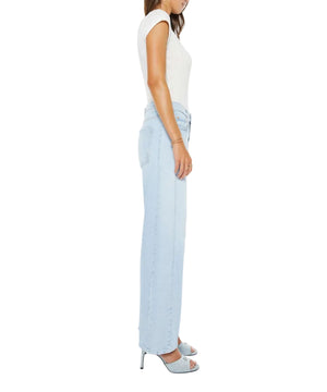 A woman in profile wears Mother’s Mid Rise Half Pipe Hover light blue, wide-leg jeans with a white short-sleeve top and open-toe heels, standing against a plain white background.