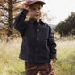 A young child wears the Rylee & Cru Baby Boys’ and Boys’ Chore Coat—a classic dark jacket with chest patch pockets—paired with a red cap, standing in a grassy field surrounded by trees and holding the brim of their hat, making it the perfect layering piece for cool outdoor adventures.