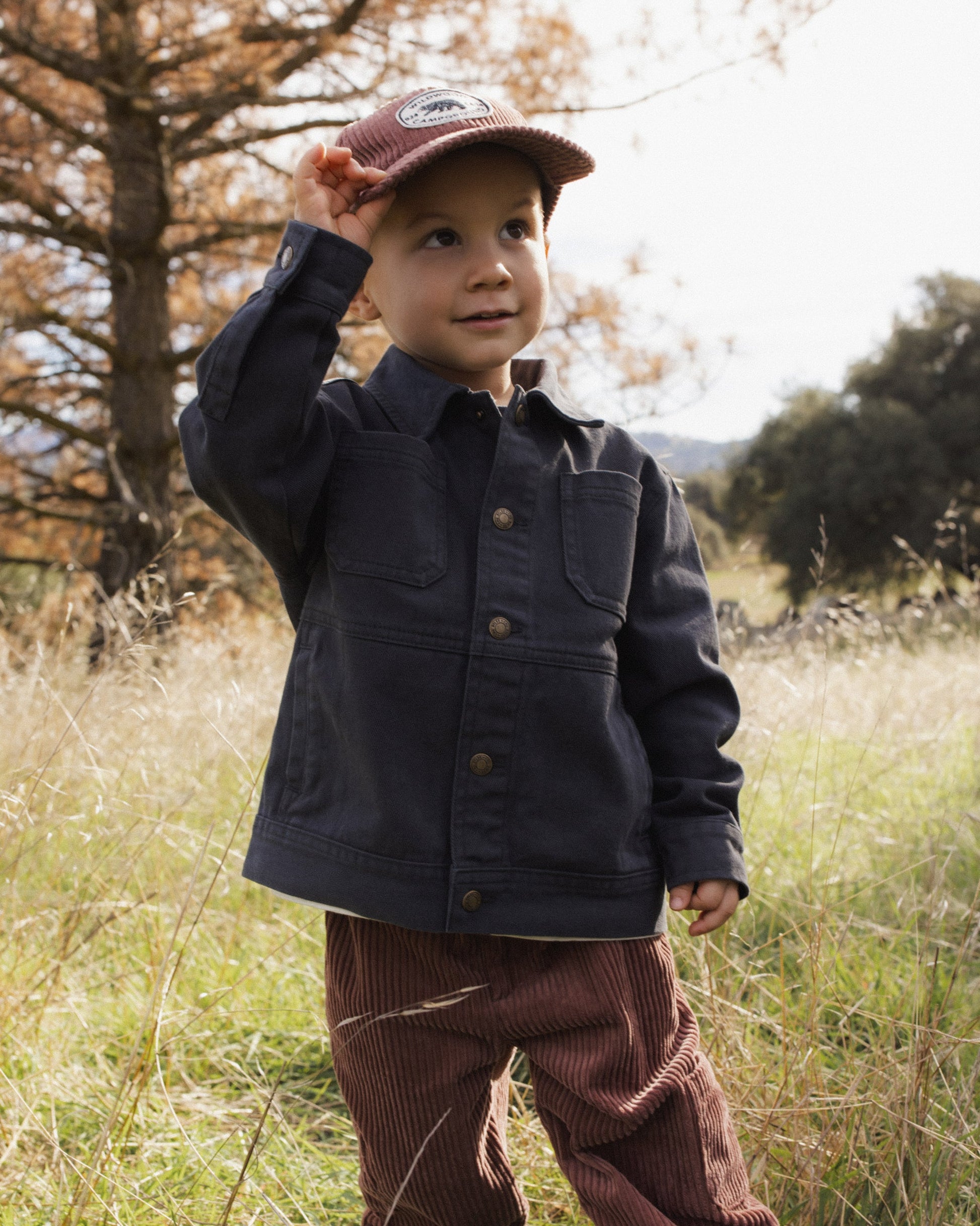 A young child wears the Rylee & Cru Baby Boys’ and Boys’ Chore Coat—a classic dark jacket with chest patch pockets—paired with a red cap, standing in a grassy field surrounded by trees and holding the brim of their hat, making it the perfect layering piece for cool outdoor adventures.