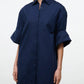 A woman wears the Staud Nico Dress—a navy blue, long-sleeve button-up mini shirtdress crafted from crisp cotton poplin with a chest pocket and rolled cuffs featuring adjustable sleeves—standing against a plain, light background.