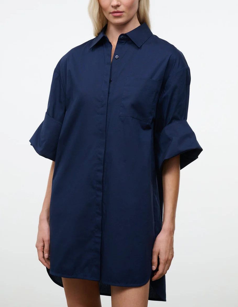 A woman wears the Staud Nico Dress—a navy blue, long-sleeve button-up mini shirtdress crafted from crisp cotton poplin with a chest pocket and rolled cuffs featuring adjustable sleeves—standing against a plain, light background.