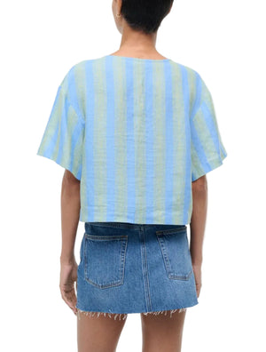 Person wearing the Staud Ava Linen Top—a short-sleeve, light blue and yellow striped linen top with adjustable front tie—paired with a frayed denim mini skirt, seen from the back.