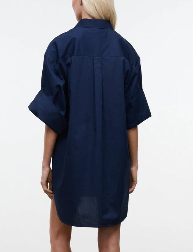 A person wearing the Staud Nico Dress—a loose-fitting, dark blue cotton poplin short-sleeve shirt dress—shown from the back against a plain, light background.