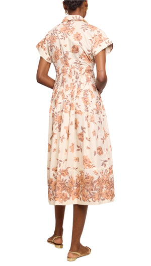 A person stands facing away, wearing the Cara Cara Nash Midi Dress—a light, fit and flare shirt dress with a brown floral pattern, pockets, and short sleeves—paired with flat sandals.
