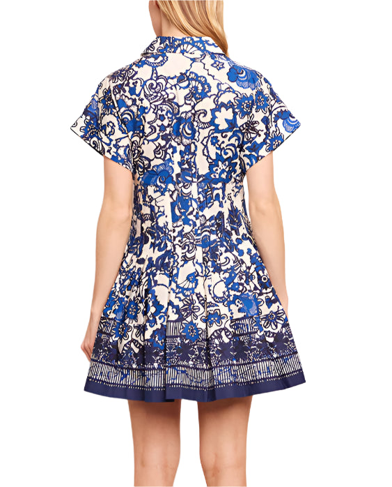 Back view of a woman in the Cara Cara Nash Mini Dress, a blue and white floral-patterned mini shirtdress with short sleeves, pintucks, and pleats.