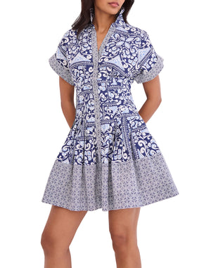 A woman wears the Cara Cara Nash Mini Dress—a fit-and-flare mini shirtdress with short sleeves, a collar, side seam pockets, and a blue and white geometric floral pattern.