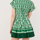 A woman stands facing away in the Cara Cara Nash Mini Dress, featuring short sleeves, a green and white geometric pattern, pleated skirt, and handy side seam pockets.
