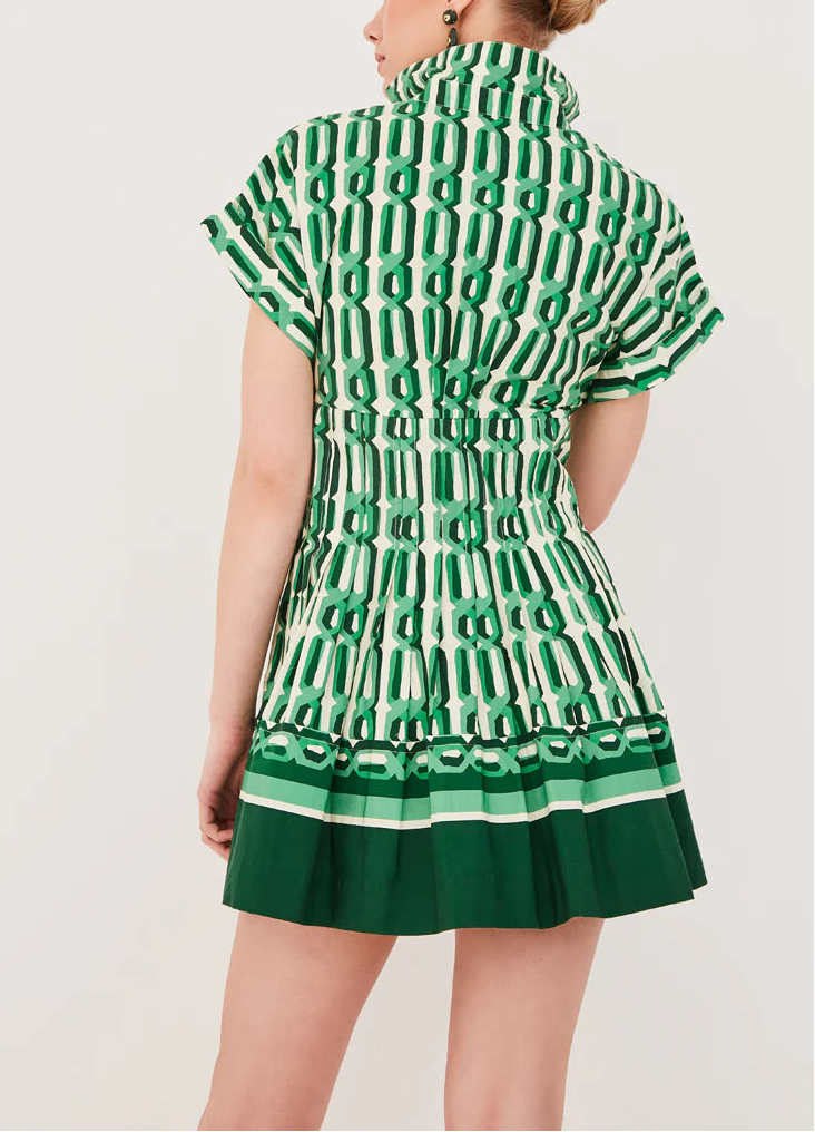 A woman stands facing away in the Cara Cara Nash Mini Dress, featuring short sleeves, a green and white geometric pattern, pleated skirt, and handy side seam pockets.