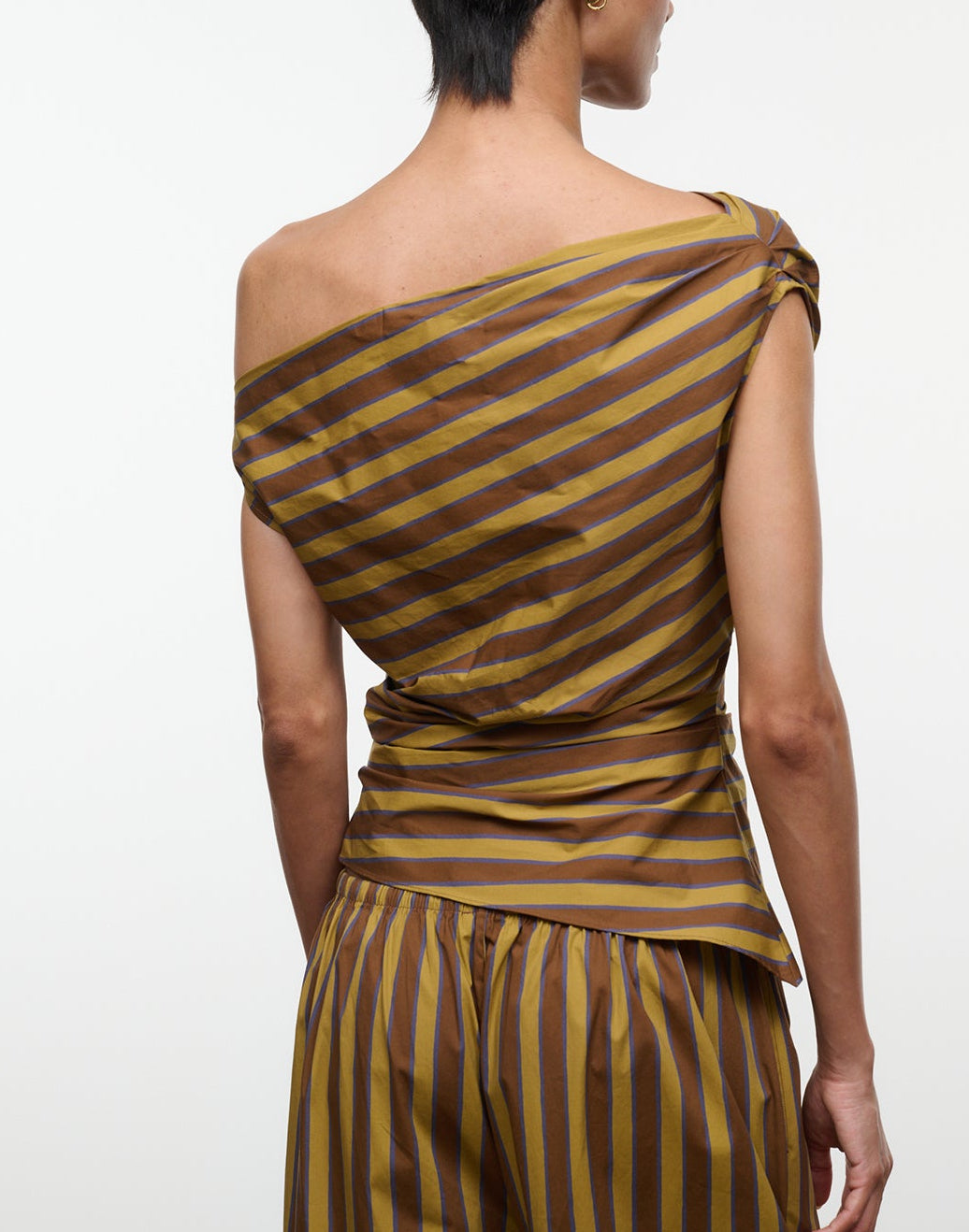 A person is photographed from the back against a plain white background, wearing the Staud Phare Top—a brown and yellow striped, off-the-shoulder top—paired with matching striped pants.