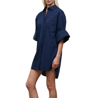 A person wearing the Staud Nico Dress, a loose-fitting navy blue cotton poplin mini shirtdress with short sleeves, stands against a plain white background.