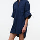 A woman is wearing the Staud Nico Dress, a loose-fitting, navy blue cotton poplin button-up mini shirtdress with adjustable sleeves, standing against a plain white background.