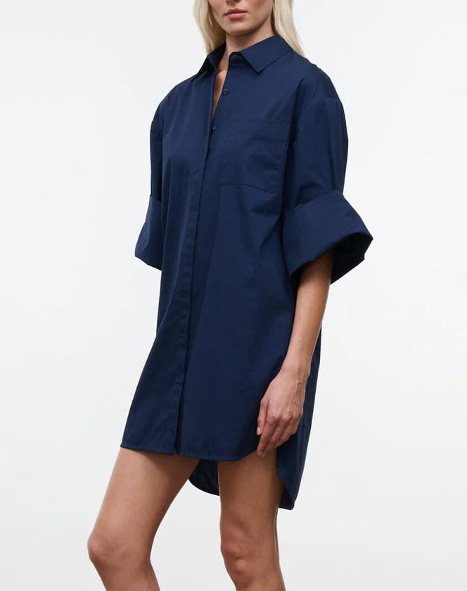 A woman is wearing the Staud Nico Dress, a loose-fitting, navy blue cotton poplin button-up mini shirtdress with adjustable sleeves, standing against a plain white background.