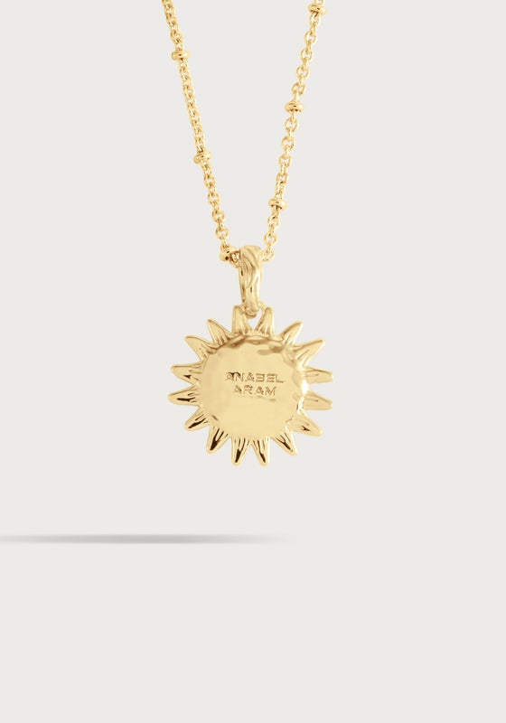 The Milos Soleil Charm by Anabel Aram is a gold sunburst necklace with a sun-shaped pendant engraved with "NASEL ARAM," inspired by Mediterranean sun jewelry, shown against a plain light background.