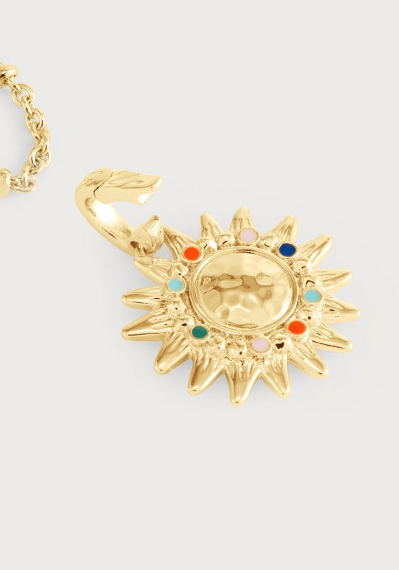 The Milos Soleil Charm by Anabel Aram is a gold sunburst necklace with a textured, sun-shaped pendant featuring colorful round stones and an attached chain, inspired by Mediterranean sun jewelry.