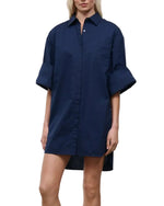 A person wearing the Staud Nico Dress, an oversized navy blue cotton poplin button-up shirt dress with adjustable sleeves, standing against a white background.