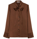 The CO Scarf Blouse is a brown, long-sleeve charmeuse top featuring a button-down front and a statement bow tie at the neck.