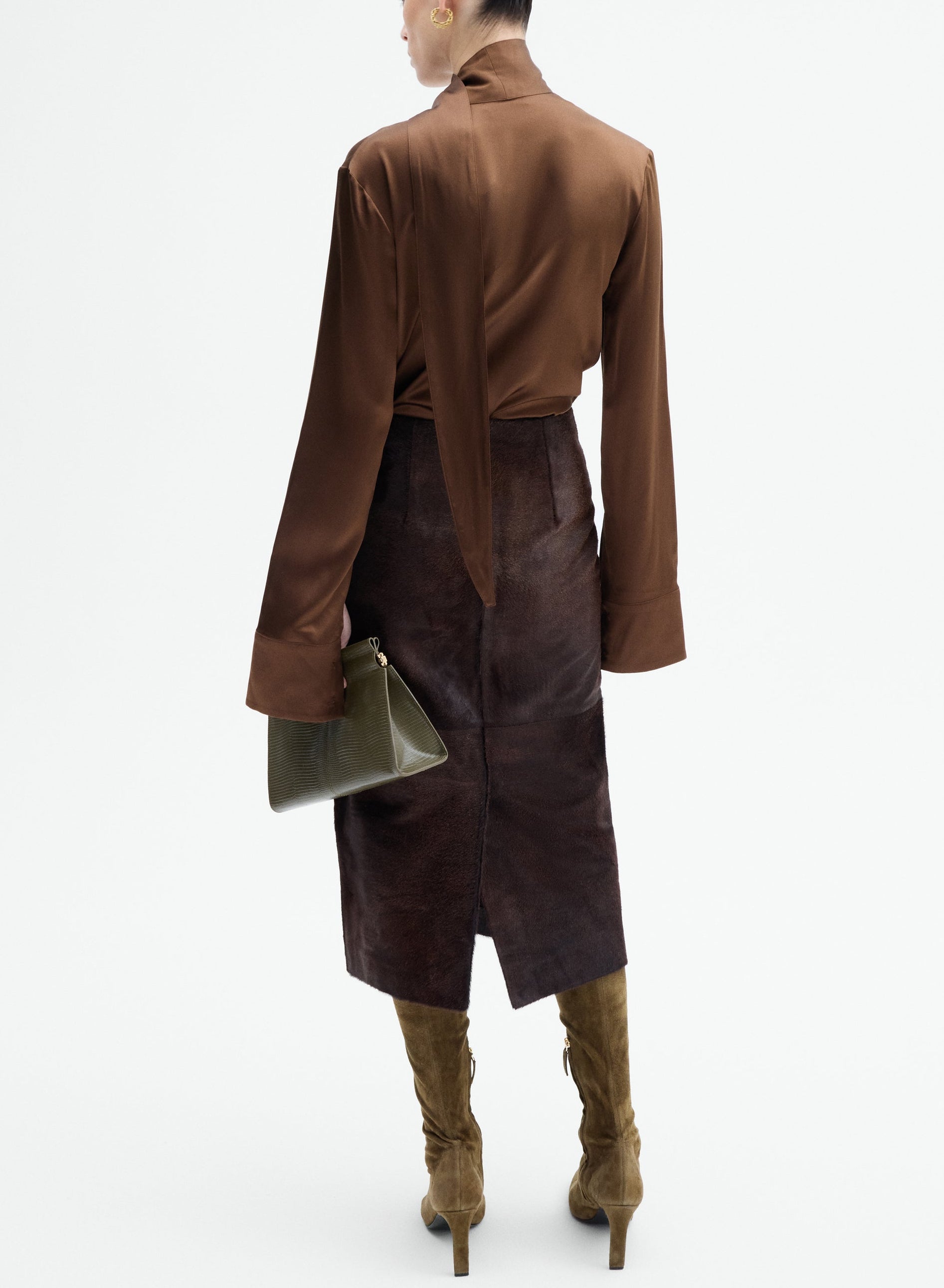 Person stands with back to camera wearing the CO Scarf Blouse in brown silk, a dark brown midi skirt, olive green knee-high boots, and holding an olive green clutch bag.