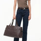A person in a brown short-sleeve top, dark blue jeans, and brown boots holds the Staud Piper Tote, a large dark brown work bag with buckle detailing, against a plain white background.