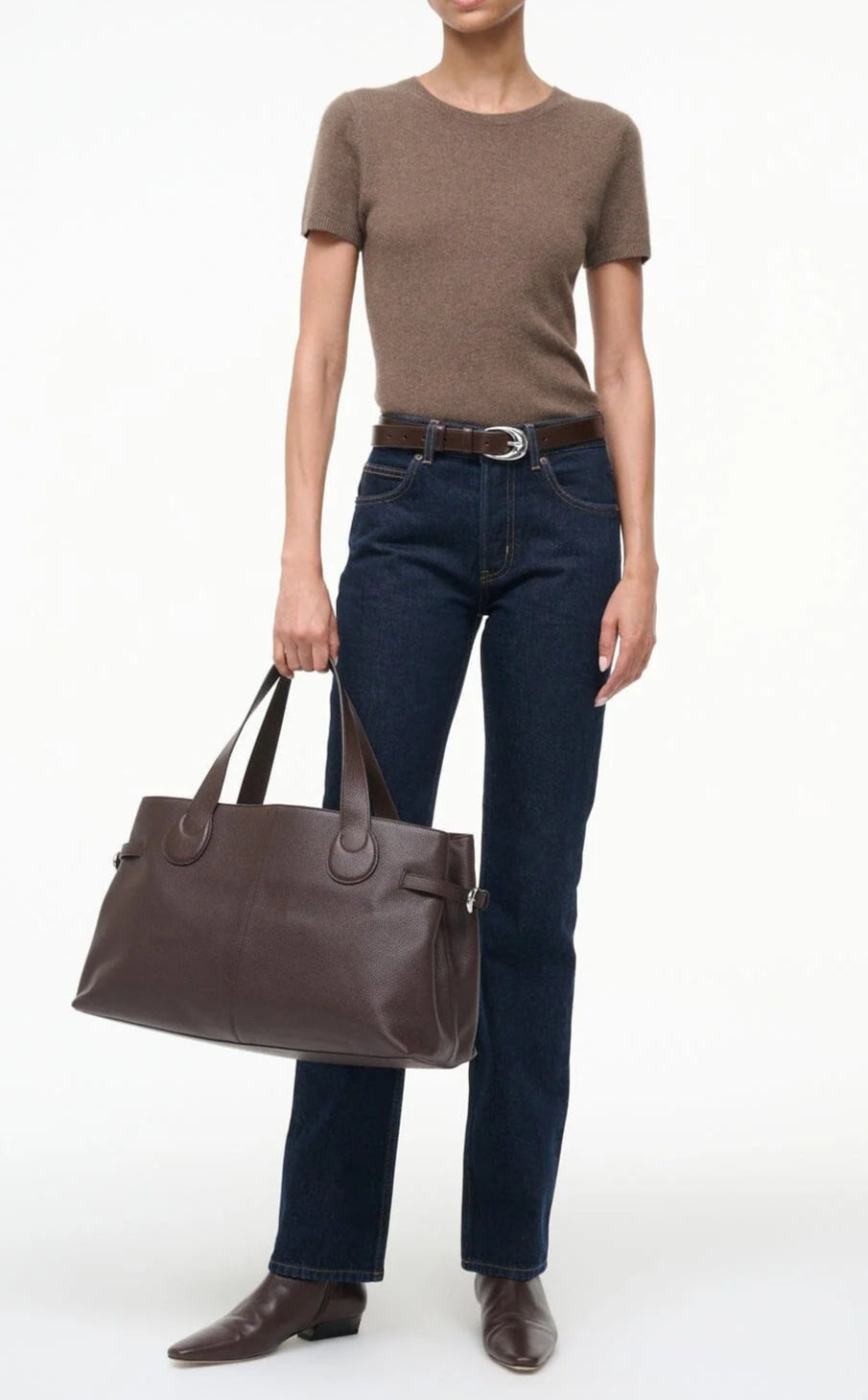 A person in a brown short-sleeve top, dark blue jeans, and brown boots holds the Staud Piper Tote, a large dark brown work bag with buckle detailing, against a plain white background.