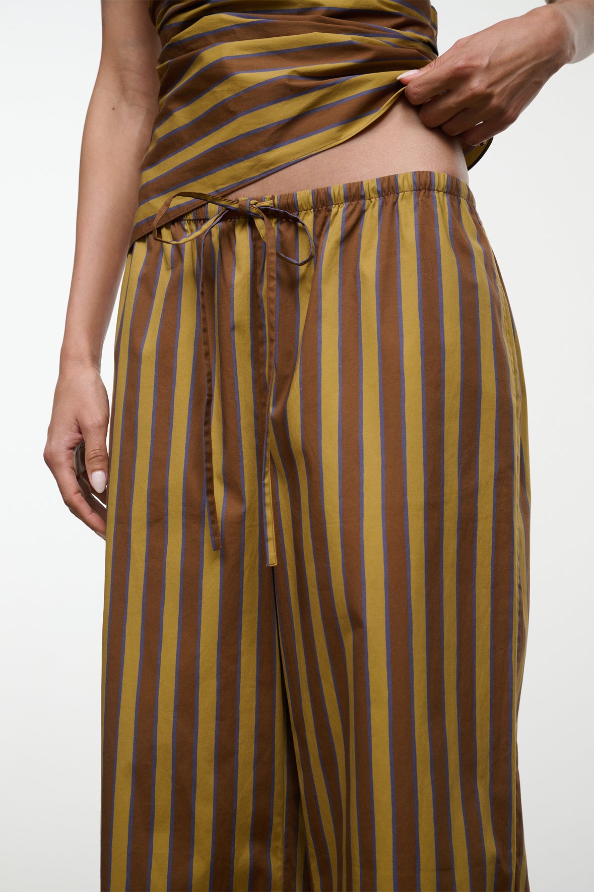 A person lifts the top of their brown and blue striped outfit to reveal the drawstring waist and waistband of Staud’s Mateo Cotton Poplin Pant.