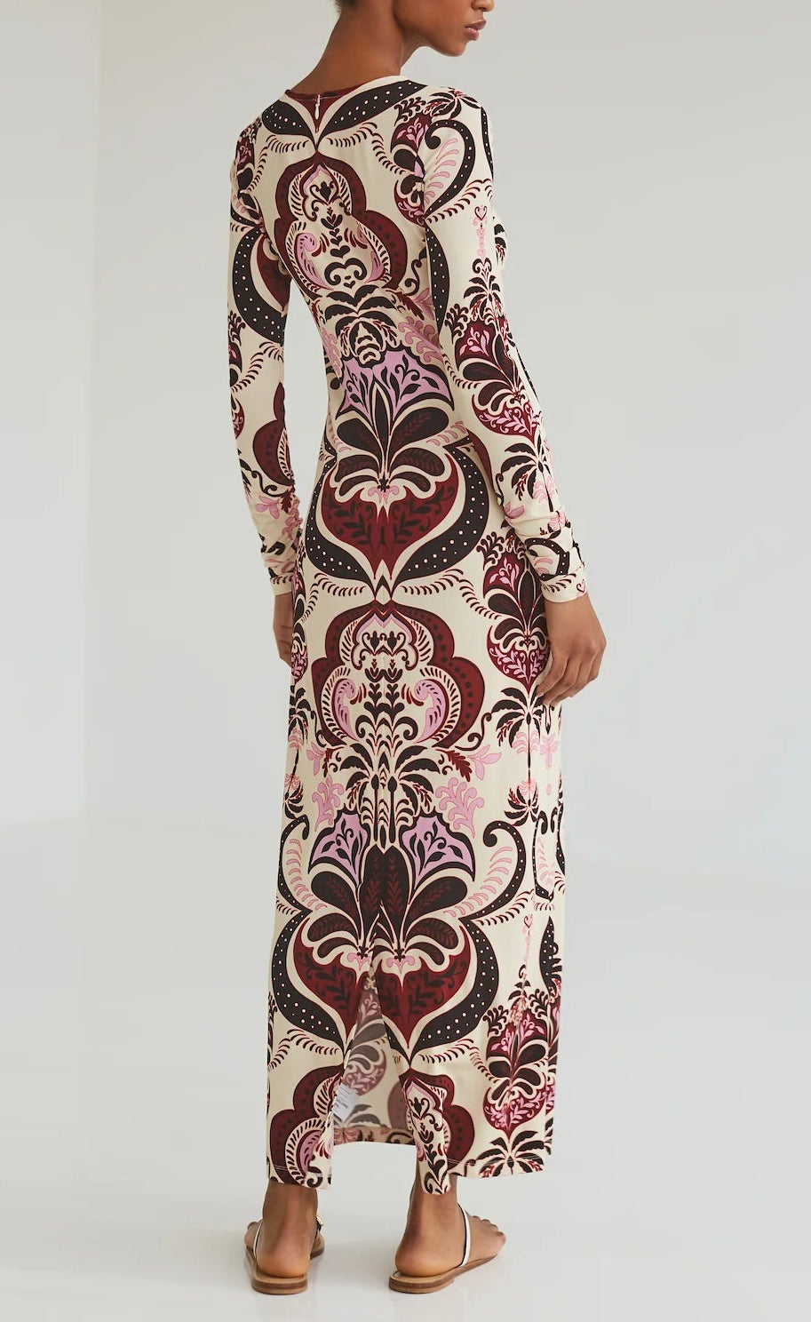 A person stands facing away, wearing the Cara Cara Danae Midi Dress—a crewneck with long sleeves and a bold maroon, pink, and cream pattern—paired with flat sandals.
